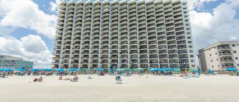 A high-rise hotel with multiple balconies, a sandy beach in front, and blue umbrellas.