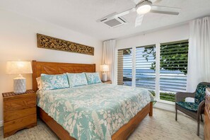 Ein Schlafzimmer mit einem hölzernen Bett, zwei Nachttischen mit Lampen, einem Deckenventilator und Meerblick durch große Fenster.