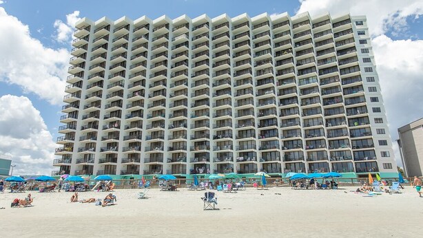 Een hoog hotel met verschillende balkons, een zandstrand ervoor en strandparasols.