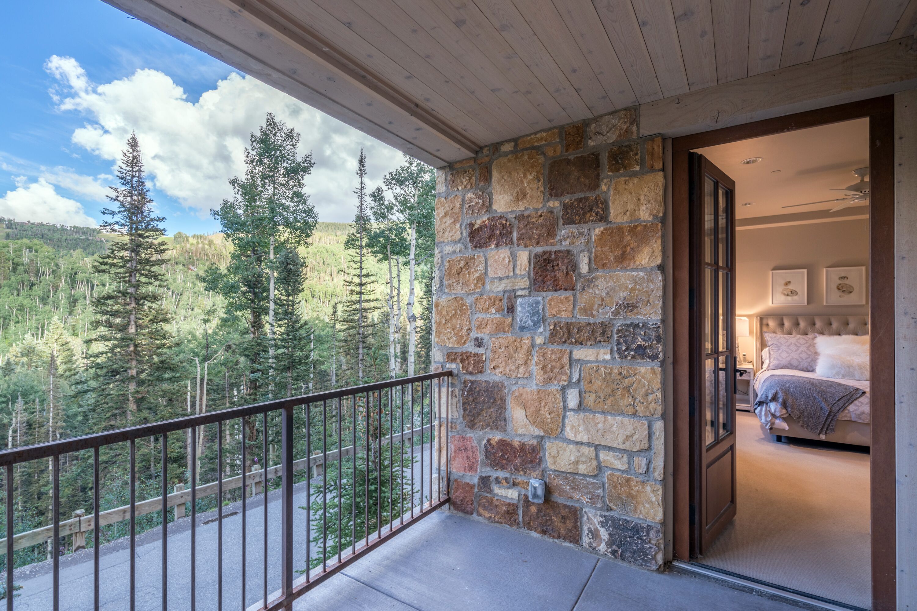 Un balcon avec un mur en pierre, une chambre à coucher avec un lit et deux tableaux encadrés, et une vue sur une zone boisée.
