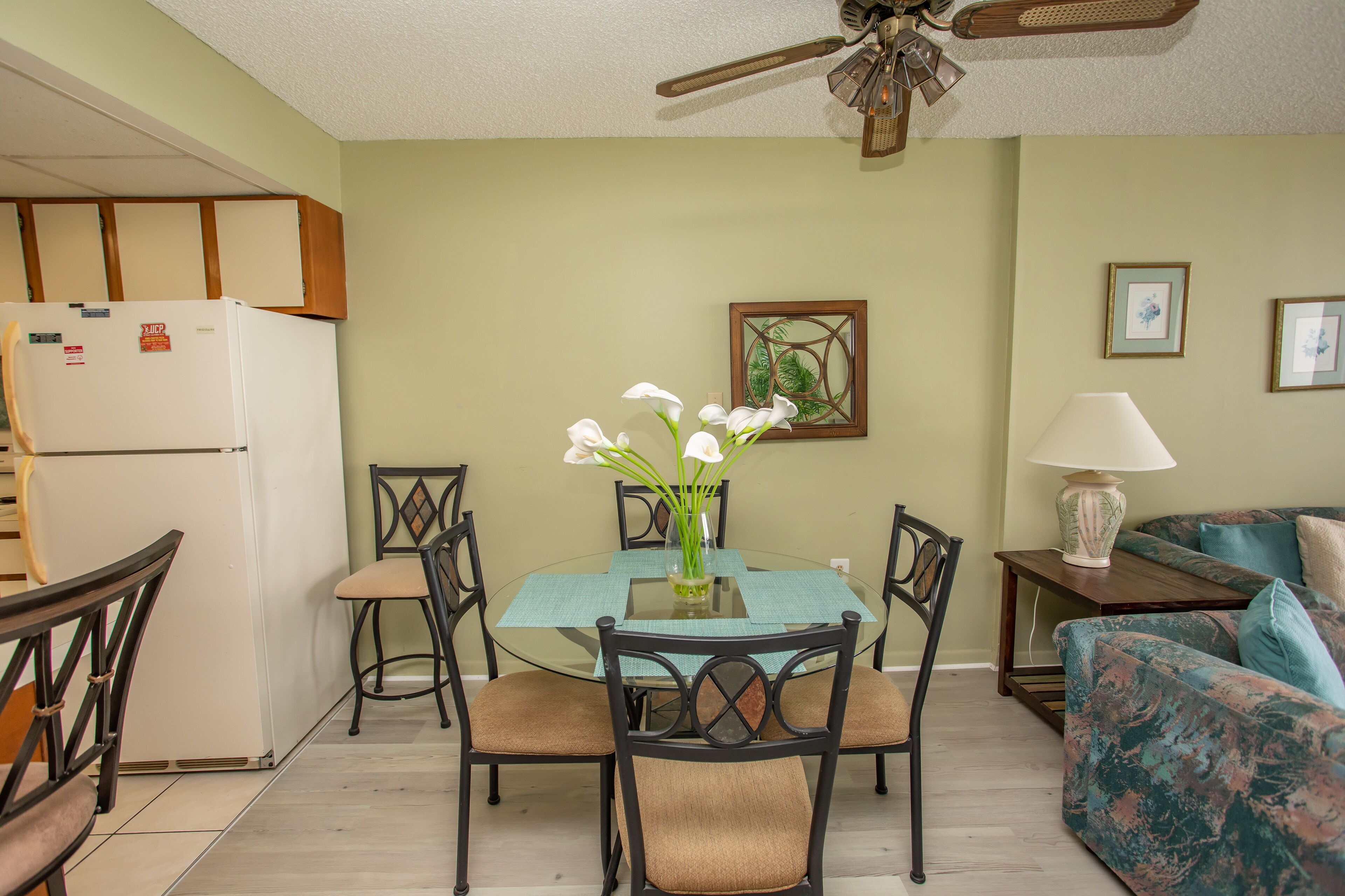 A dining area with a glass table surrounded by chairs, a refrigerator, a ceiling fan, and a couch.