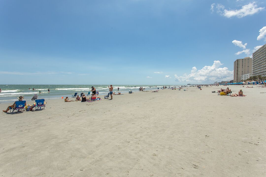 A beach with people sunbathing and swimming, a pier in the background, and a large hotel building.