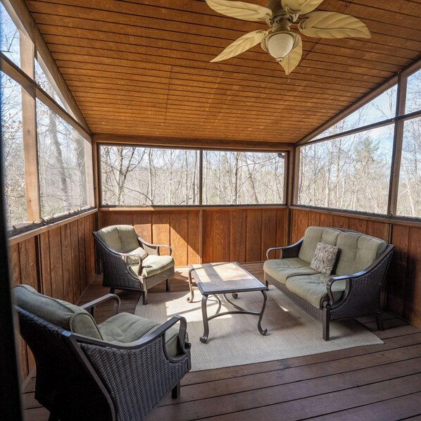 A covered outdoor seating area with wicker furniture, a ceiling fan, and large windows offering a view of the surrounding trees.