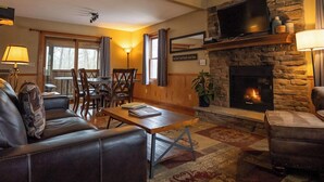 A cozy living room with a fireplace, a television mounted on a stone wall, and a dining area with wooden furniture.