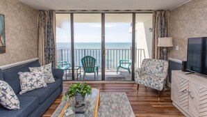 A living room with a sofa, armchair, and coffee table, a TV, and a balcony with chairs and a view of the sea. - 14th-floor oceanfront condo with breathtaking views (Murrells Inlet)