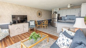 A modern living room with a sofa, coffee table, and TV. There is a dining area with chairs and a table. The kitchen is visible with cabinets and appliances. - 14th-floor oceanfront condo with breathtaking views (Murrells Inlet)