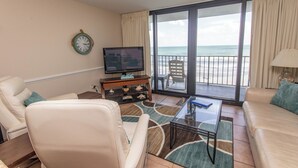Sala de estar com televisão, dois sofás, mesa de centro e vista para o mar através de grandes portas deslizantes.