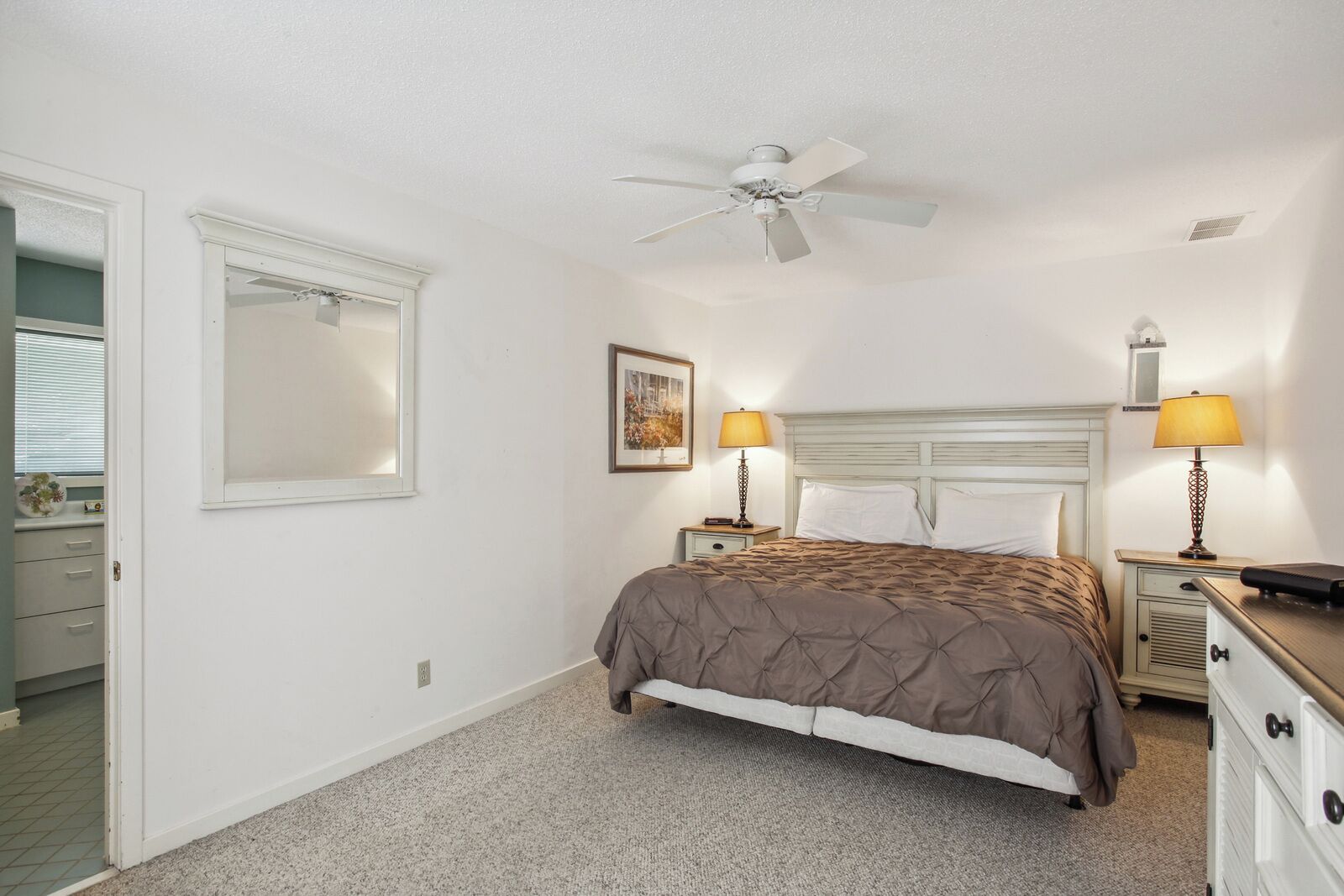 Une chambre à coucher avec un lit, deux tables de chevet avec des lampes, un ventilateur de plafond, un tableau encadré au mur et une porte donnant sur une autre pièce.