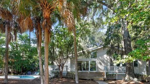 A house with a wooden deck, surrounded by trees and a pathway leading to a pool.