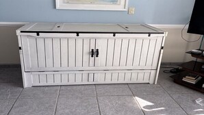 Uma unidade de armazenamento branca com duas portas e parte superior plana, colocada em um quarto com paredes azul-claras e um quadro pendurado acima.