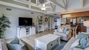 Amplia sala de estar con televisor grande, ventilador de techo y una zona de cocina con armarios de madera y una mesa de comedor.