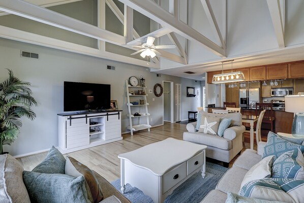 Amplia sala de estar con televisor grande, ventilador de techo y una zona de cocina con armarios de madera y una mesa de comedor.
