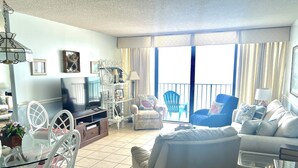 A spacious living room with a television, white furniture, a balcony with chairs, and a large window with curtains. - LARGE AND LOVELY! LOTS OF ROOM IN THIS 3 BEDROOM CONDO RIGHT ON THE OCEAN! (Murrells Inlet)