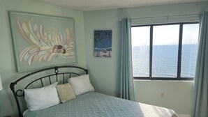A bedroom with a bed, a nightstand, a painting, and a window with a view of the sea. - LARGE AND LOVELY! LOTS OF ROOM IN THIS 3 BEDROOM CONDO RIGHT ON THE OCEAN! (Murrells Inlet)
