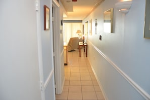 A narrow hallway with a chair and a lamp in the distance, leading to a room with a bed.