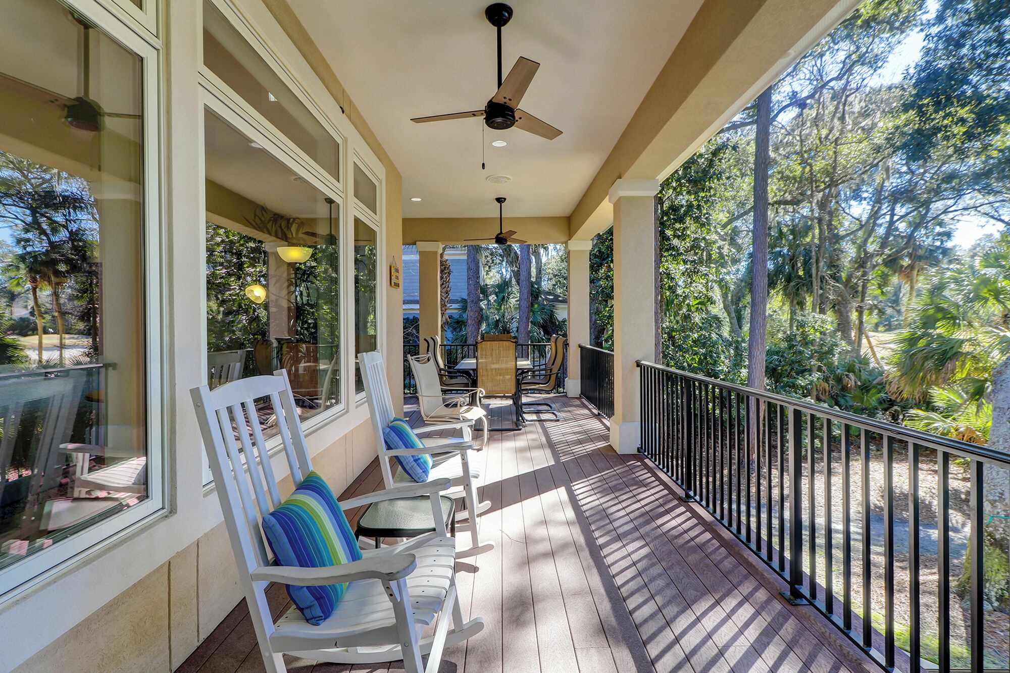 A covered outdoor area with a ceiling fan, white chairs with cushions, and a view of trees and greenery.