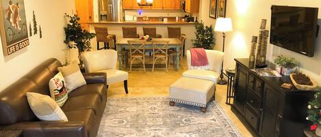 A living room with a brown leather sofa, a white ottoman, and a patterned rug. There is a dining area with a wooden table and chairs, and a kitchen in the background with wooden cabinets and a refrigerator.