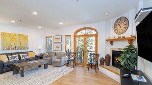 A spacious living room with a fireplace, a large clock, and a view of the outdoors.