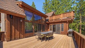 A wooden deck with a dining table and chairs, large windows, and a deck railing.