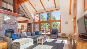 A spacious living room with a stone fireplace, large windows, a wooden staircase, and a cozy seating area.