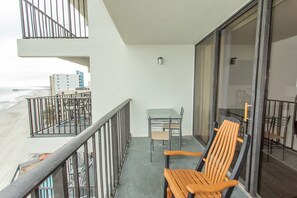 Een balkon met een houten stoel, een glazen tafel en uitzicht op het strand en gebouwen.