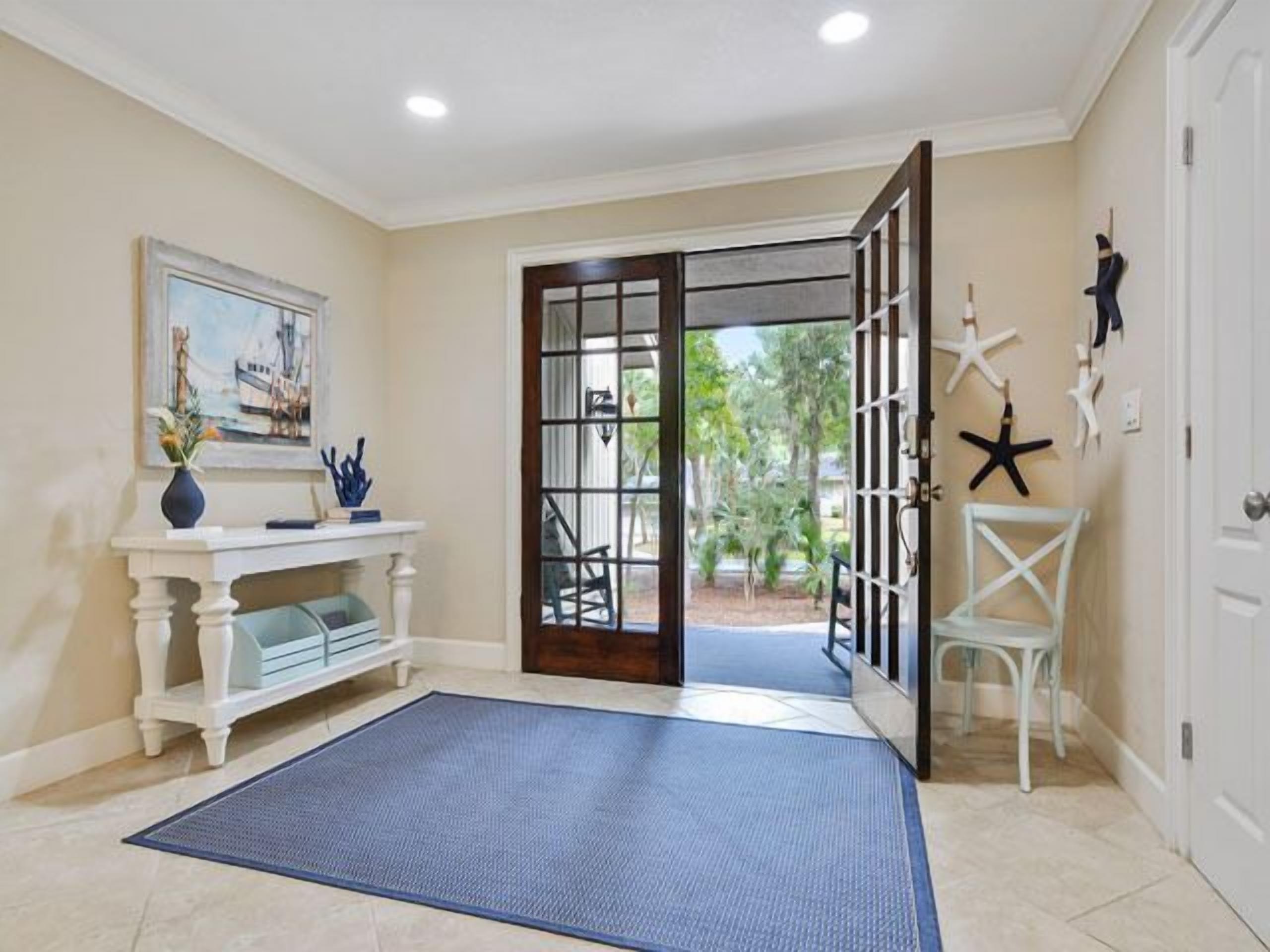 Une pièce avec une console blanche, un tableau, un tapis bleu, une porte à panneaux de verre et des décorations en forme d’étoiles de mer.