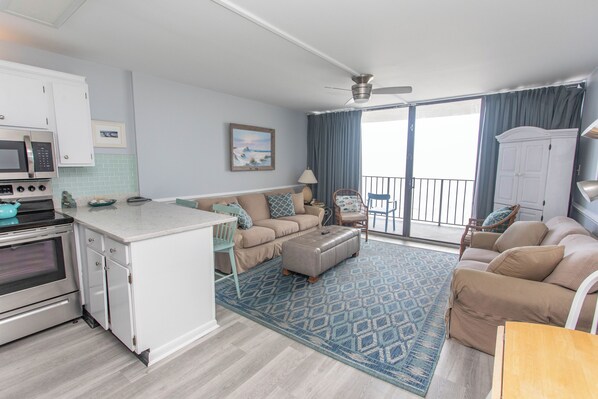 A modern kitchen with white cabinets, a microwave, and an oven. There is a dining area with a table and chairs, a sofa, and a patterned rug. A balcony with a view is visible through the glass doors. - Beachfront Relaxation Starts Here (Murrells Inlet)