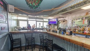 A bar area with a wooden counter, black bar stools, a television screen, and a variety of bottles. - OCEANFRONT LARGE 2 BEDROOM CONDO (Murrells Inlet)