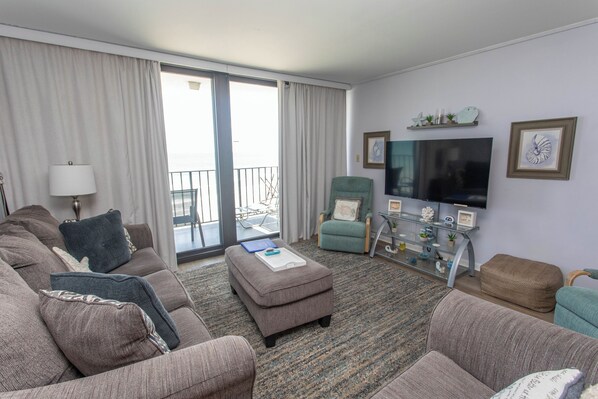 A living room with a sofa set, ottoman, and armchairs, a flat-screen TV, a glass coffee table, and a balcony with a view. - OCEANFRONT LARGE 2 BEDROOM CONDO (Murrells Inlet)