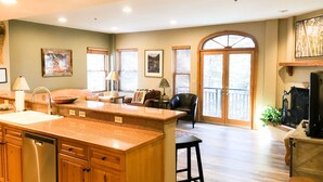 A spacious kitchen with a large island, wooden cabinets, and a dishwasher. There is a dining area with a chair and a table, and a large window with blinds.