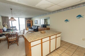 A spacious living area with a kitchen island, dining table, and chairs. There is a balcony with a view, a sofa, and a television.