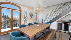 Un comedor con una mesa de madera y sillas azules, una araña, una escalera y vistas de montañas nevadas.