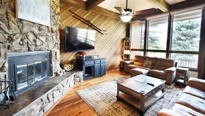 A cozy living room with a stone fireplace, a television, a leather sofa, and large windows offering a view of trees. - Near Lake Dillon! 3 Kings. Hot Tub, Yard. On Rec Path, Near Dining, Shops (Keystone)
