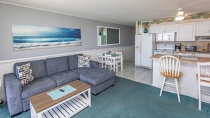 A compact living space with a grey sofa, a wooden coffee table, a dining area with a white table and chairs, and a kitchen with white cabinets and appliances. - SEA WORTHY 2 BEDROOM OCEAN FRONT CONDO (GARDEN CITY BEACH)