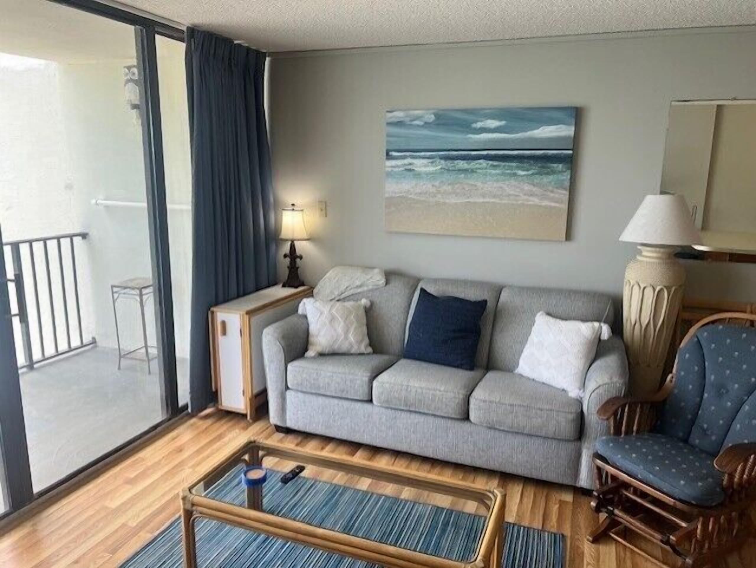 A living room with a grey sofa, a wooden coffee table, a blue armchair, a painting of a beach, and a balcony with a view.