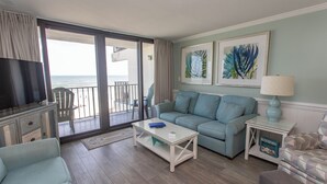 Ein Wohnzimmer mit einer Couch, einem Couchtisch und einem Fernseher. Es gibt einen Balkon mit Meerblick.