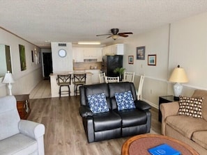A living room with a black leather sofa, a beige sofa, a wooden coffee table, and a dining area with a kitchen in the background.