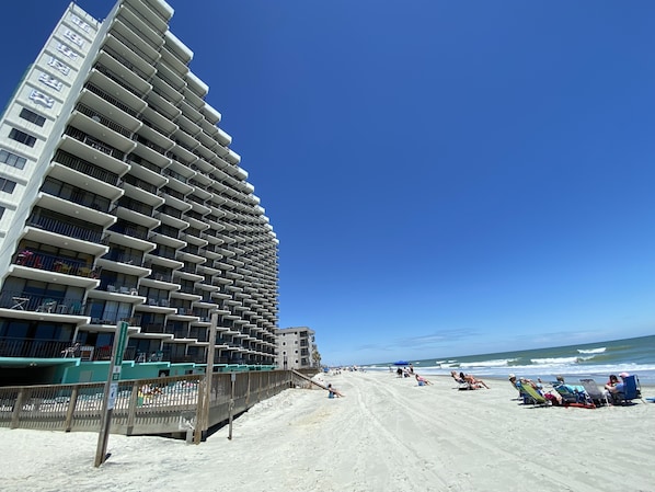 Un hôtel de grande hauteur dot é de nombreux balcons, une plage de sable au premier plan et l’océan en arrière-plan.