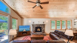 A spacious living room with a stone fireplace, leather sofas, a wooden ceiling fan, and large windows offering a view of the outdoors.
