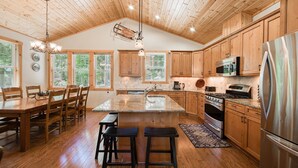 A spacious kitchen with wooden cabinets, a large island with stools, a stainless steel refrigerator, and a wooden ceiling.