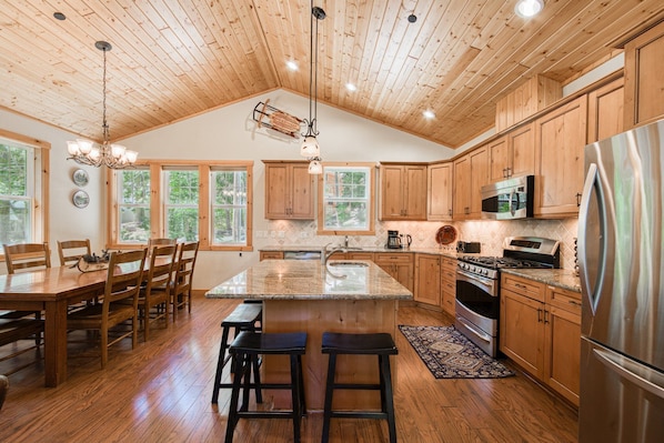 A spacious kitchen with wooden cabinets, a large island with stools, a stainless steel refrigerator, and a wooden ceiling.