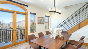 A dining area with a large wooden table, multiple chairs, a chandelier, and a view of mountains through a large window.