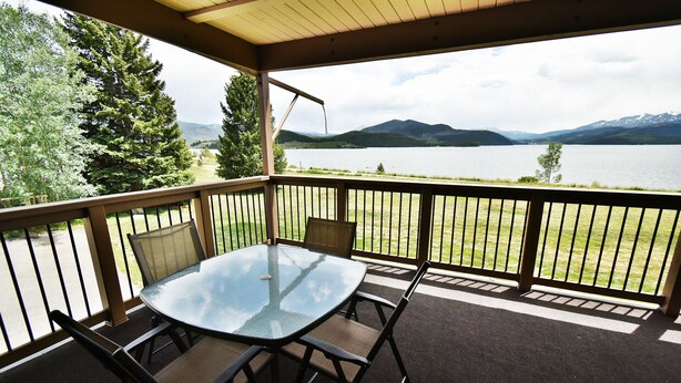 A covered outdoor area with a glass table and chairs, a view of a lake and mountains, and a wooden railing. - On Lake, Views! Walk to Amphitheater, Marina, Dining. End Unit, Deck (Dillon)