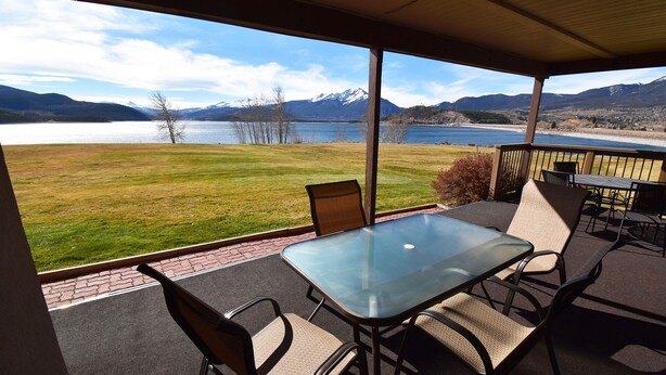 A covered patio with a glass table and chairs, overlooking a lake and mountains. - On Lake! Views, 1st Floor, Hot Tub! Walk to Amphitheater, Marina, Dining (Dillon)