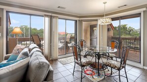 Een ruim balkon met een glazen deur die uitkomt op een terras, ingericht met een glazen tafel en stoelen, en een bank met kussens.