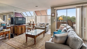 Een ruime woonkamer met een televisie, bank en een balkon met buitenmeubels.