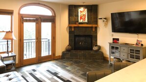 A living room with a fireplace, a television, and a stone floor. - 2-Bedroom With Mountainside Balcony, Hot Tub & Pool Access (Mountain Village)