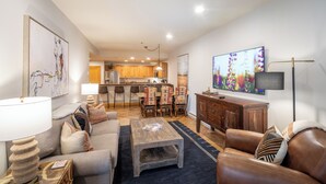 A modern living room with a sofa, coffee table, and a TV. There is a kitchen in the background with wooden cabinets and a dining area with chairs. - Mountainside 2-Bedroom Condo W/Hot Tub, Fireplace, Ski Access (Mountain Village)