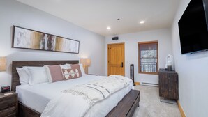 A bedroom with a large bed, wooden headboard, bedside tables, a nightstand, a wall-mounted TV, a wooden door, and a window with blinds. - Mountainside 2-Bedroom Condo W/Hot Tub, Fireplace, Ski Access (Mountain Village)