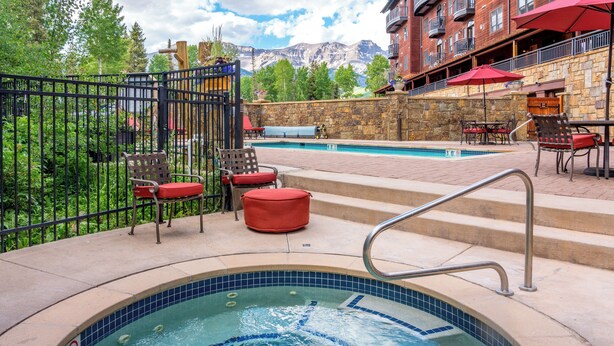 A hot tub with a metal ladder, surrounded by outdoor seating with red cushions, a stone wall, and a view of mountains in the background. - Mountainside 2-Bedroom Condo W/Hot Tub, Fireplace, Ski Access (Mountain Village)